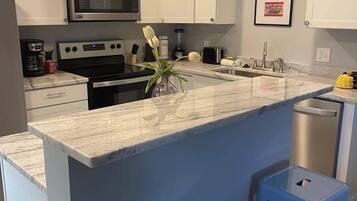 A modern kitchen with white cabinets, a stainless steel microwave, a built-in oven, a sink, and two blue stools.