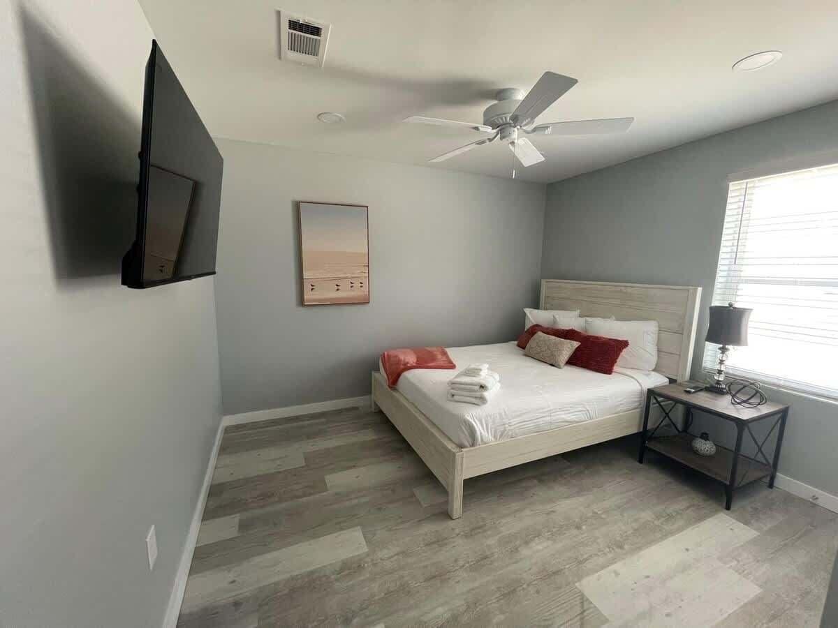 A bedroom with a bed, a nightstand, a ceiling fan, a wall-mounted TV, and a window.