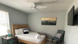 Un dormitorio con cama, una silla, una mesita de noche, un ventilador de techo y un cuadro en la pared.