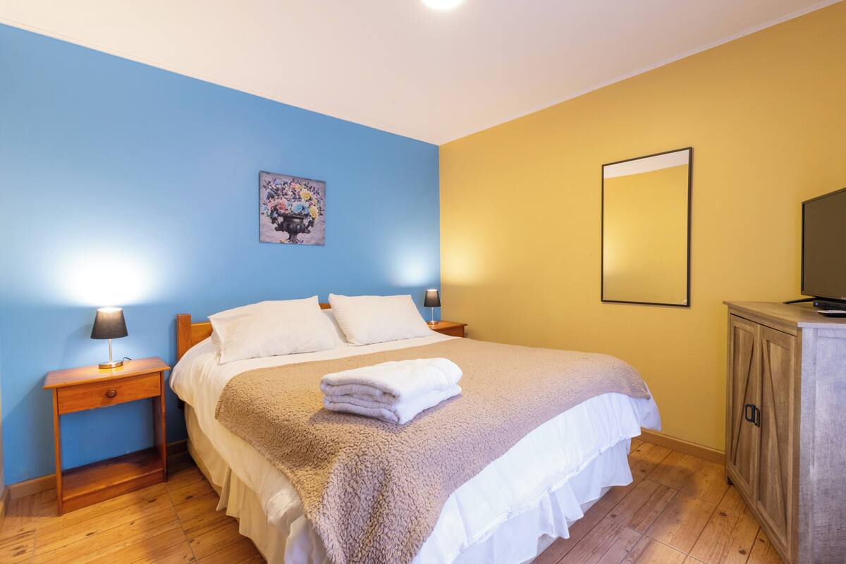 Une chambre à coucher avec un lit, des tables de chevet, une lampe, un tableau, un miroir et un téléviseur posé sur un meuble.
