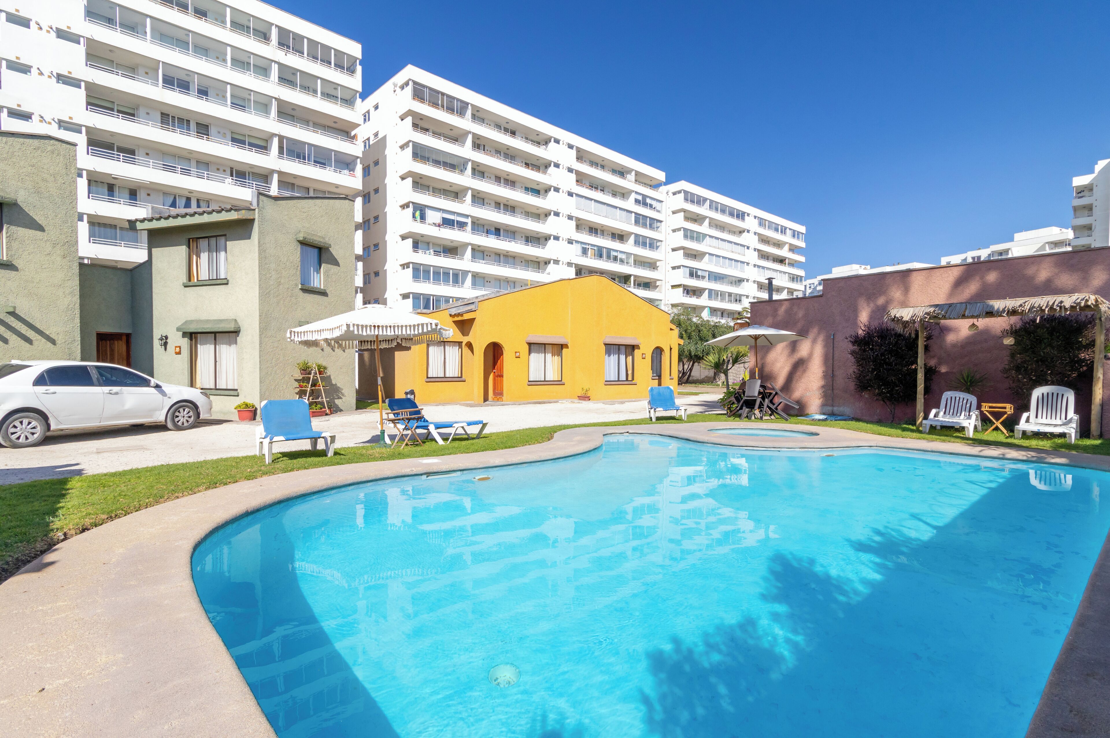 A swimming pool with lounge chairs, a yellow building, and a white car parked nearby.