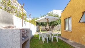 A patio with a table and chairs, a barbecue grill, and a white wall with a window. - Cabañas de la Costa (La Serena)