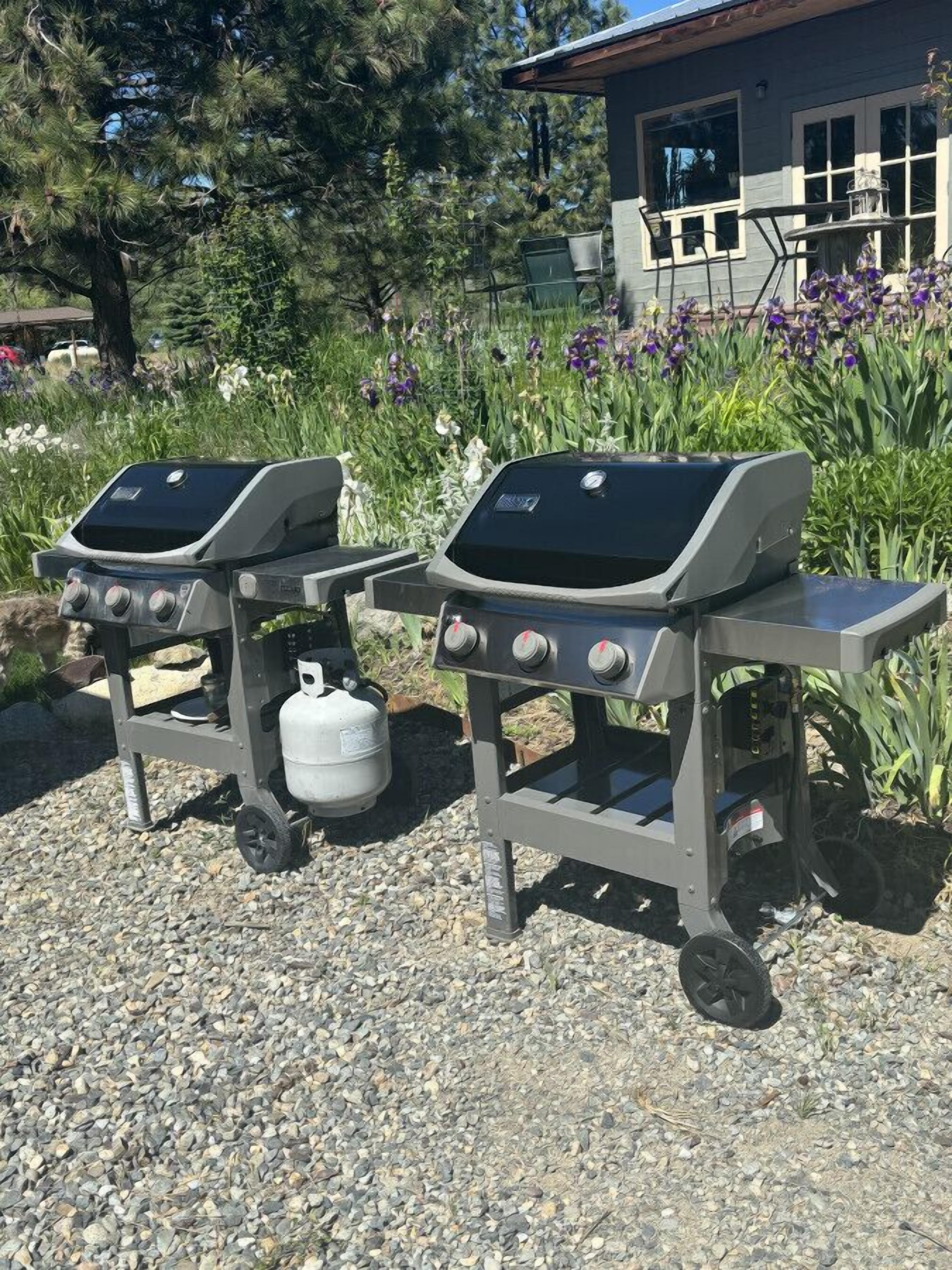 Two portable gas grills on a gravel surface with a house and garden in the background.