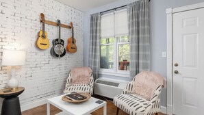 A room with a white brick wall, two patterned chairs, a white table with a book and a basket, a lamp, and a window with curtains. - Gypsy Rose: Free Parking and Steps to Downtown Charm (Eureka Springs)