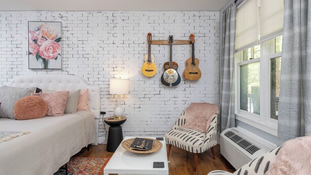 A cozy living room with a white brick wall, a white sofa, a patterned armchair, a small side table with a lamp, and a window with curtains. - Gypsy Rose: Free Parking and Steps to Downtown Charm (Eureka Springs)