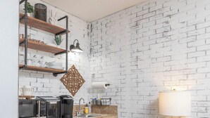 A compact kitchen with a white brick wall, wooden shelves, a black countertop, and a black stool. - Gypsy Rose: Free Parking and Steps to Downtown Charm (Eureka Springs)