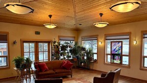 A spacious living room with wooden floors, a brown leather sofa, a red armchair, and a coffee table. There are large windows with blinds, a staircase, and a wall-mounted shelf with plants.