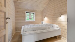 Un dormitorio con techo inclinado de madera, una cama individual con ropa de cama blanca, una ventana con vistas a zonas verdes y una lámpara de noche.