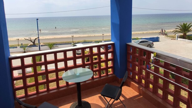A balcony with a glass table and chairs, overlooking a beach with a clear view of the sea and a road with vehicles.