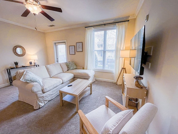 A living room with a sofa, coffee table, and armchair, a ceiling fan, and a television on a stand.