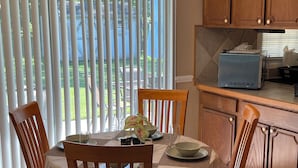 Un comedor con una mesa dispuesta para cuatro personas, rodeada de sillas de madera, una zona de cocina con armarios de madera y vistas a través de ventanas grandes con persianas.