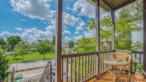 A balcony with a wicker chair, wooden railing, and a view of a green park and buildings. - Sunny, 2 Bedroom Apt near Johns Hopkins, Charles Village - Explore Baltimore! (Maryland)