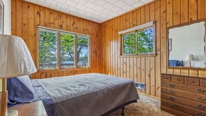 A bedroom with wooden walls, a bed, a dresser, and two windows.