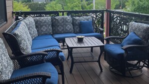 A covered outdoor seating area with a blue sofa and wicker chairs, a small table, and a view of trees and power lines. - TC West Side Condo, Central Location, Spacious, Dog Friendly! (Traverse City)