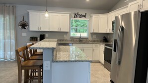 Une cuisine moderne avec des armoires blanches, un comptoir en granit, des appareils en acier inoxydable et un panneau décoratif portant l’inscription « Bénite » au mur.