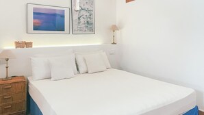 Ein ordentlich bezogenes Bett mit weißen Leinen und Kissen, ein Nachttisch mit Lampe sowie zwei gerahmte Kunstwerke an der Wand.