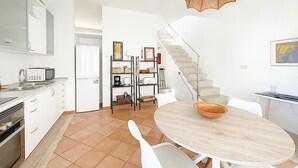 Eine moderne Küche mit weißen Schränken, einem eingebauten Backofen und einer Mikrowelle. Es gibt eine Essecke mit einem runden Tisch und Stühlen, sowie eine Treppe, die zu einer anderen Ebene führt.
