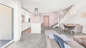 Un salon moderne avec un canapé gris, un coin repas avec une table et des chaises, une cuisine avec des meubles blancs et un escalier menant à un autre niveau.
