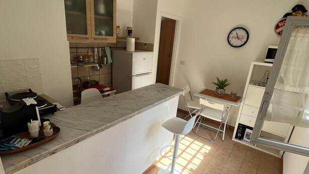 A compact kitchen with a bar area, a dining table with chairs, a refrigerator, and a clock on the wall.