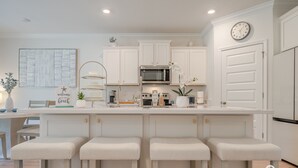 A modern kitchen with white cabinetry, a central island with bar stools, a microwave, and a clock on the wall. - Elegant Home 4BR/2BA Minutes From 30A & Sandestin Shopping (Santa Rosa Beach)