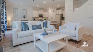 A modern living room with a white sofa, coffee table, and decorative items. A kitchen is visible in the background with white cabinets and appliances. - Elegant Home 4BR/2BA Minutes From 30A & Sandestin Shopping (Santa Rosa Beach)