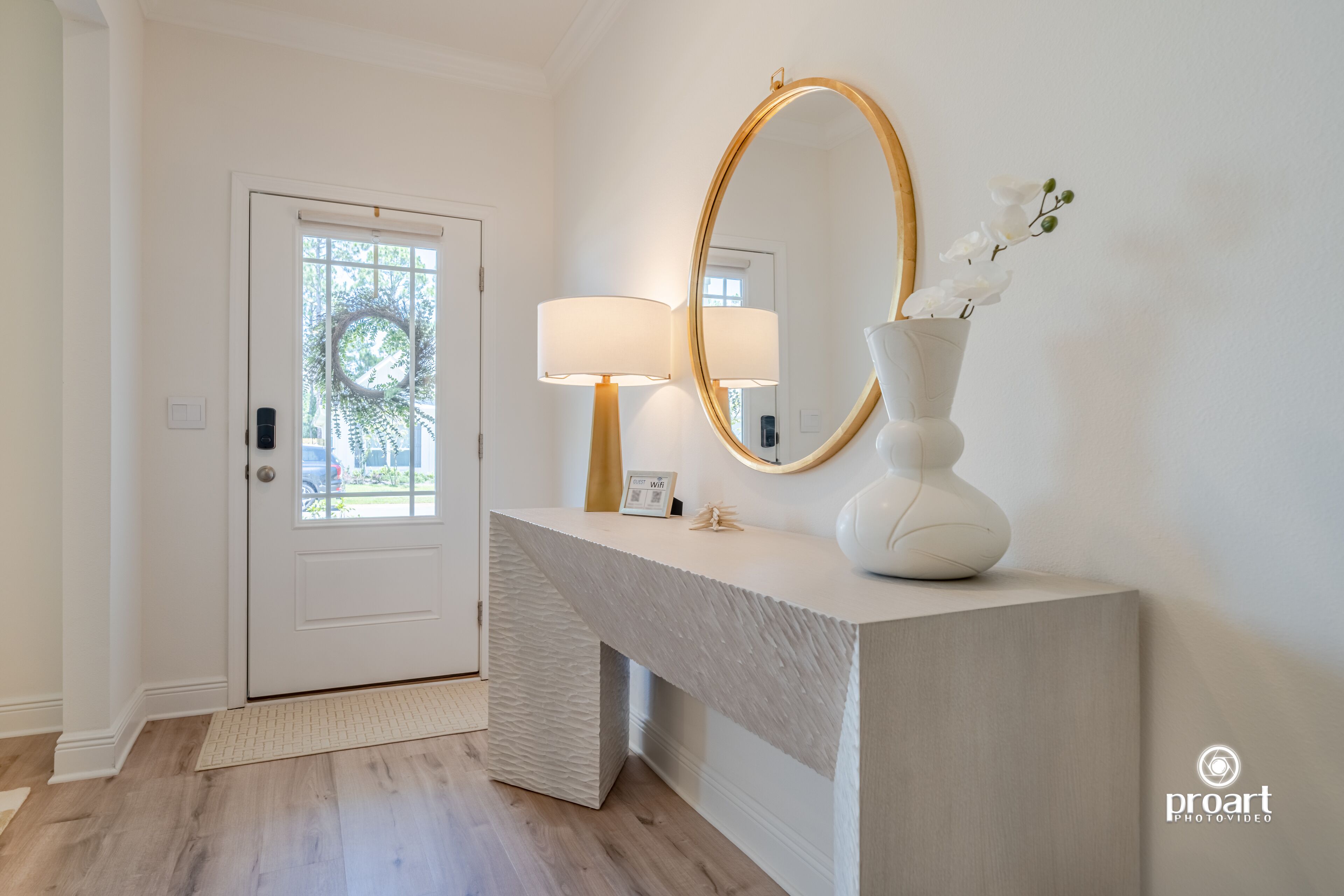 A modern entryway with a white door, a textured console table, a wall-mounted mirror, and a vase with white flowers.