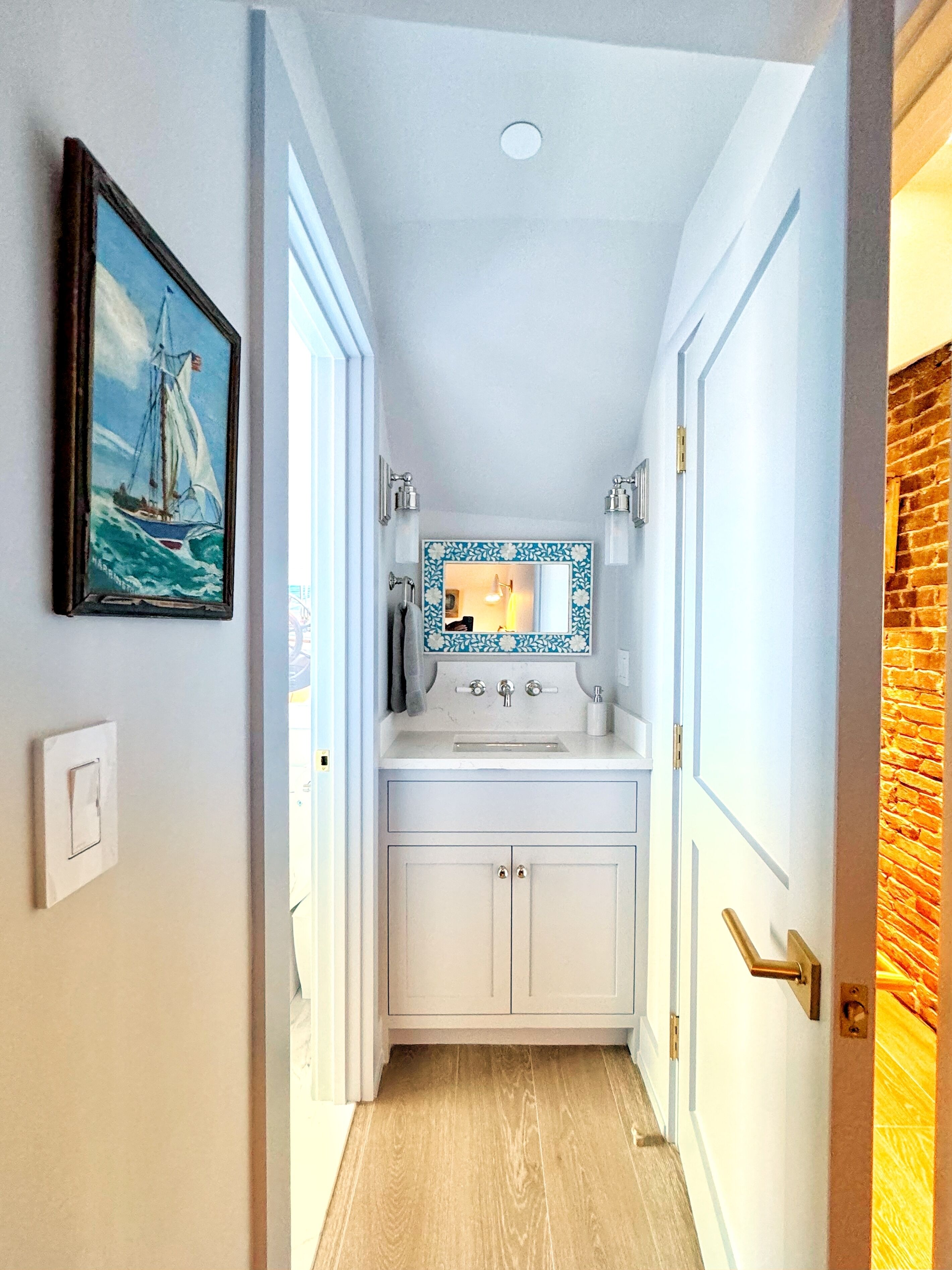 A narrow hallway leading to a bathroom with a white sink, a mirror with a floral frame, and a painting of a sailboat on the wall.