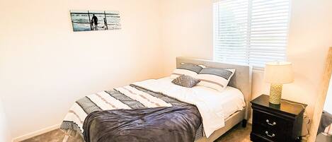 A bedroom with a bed, a nightstand, a lamp, a window with blinds, and a wall art piece.