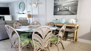 Un comedor con una mesa redonda de cristal, sillas de mimbre con cojines verdes, una araña, un cuadro de un muelle y un aparador de madera con adornos.