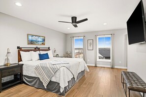 A bedroom with a bed, bedside table, lamp, ceiling fan, and a TV mounted on the wall.