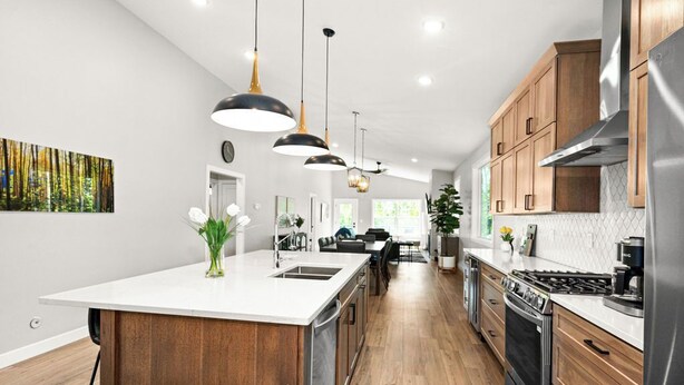 Une cuisine moderne avec des meubles en bois, un îlot central, des appareils en acier inoxydable, un éclairage suspendu et un coin repas à l’arrière-plan.