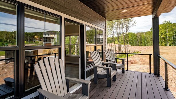 A covered outdoor deck with Adirondack chairs, large windows, and a view of a forested area.
