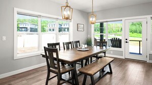 A dining room with a wooden table, chairs, and benches. There are two pendant lights hanging above the table. The room has large windows with a view of houses and greenery outside.