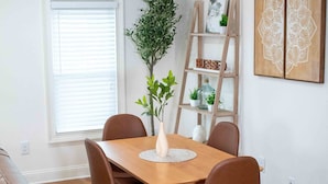 Un comedor con una mesa de madera, sillas marrones, un florero con plantas, una estantería escalonada con adornos y una ventana con persianas.