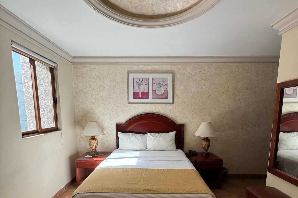 Quarto de hotel com uma cama grande, duas mesas de cabeceira com luminárias, uma luz no teto, uma janela e uma obra de arte na parede.