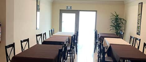 Un salón de conferencias con varias mesas y sillas, una puerta con letrero de salida de emergencia y una planta en maceta.