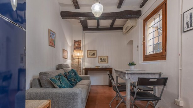 Ein kompakter Wohnbereich mit einer Couch, einem Esstisch mit Stühlen, einem kleinen Holzschemel, einem Deckenventilator und einem Fenster mit Blick.