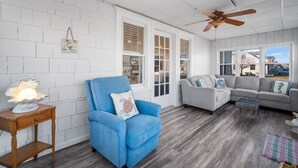A living room with a blue armchair, grey sofa, wooden flooring, white walls, and a ceiling fan.