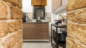 A kitchen with a brick wall, a wooden cabinet, a window with a curtain, and a small table with a kettle. - GuestHost - The Arches of Ballarò - Cortile Scimeca Apartment (Palermo)