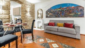 A dining area with a glass table, chairs, a stone wall, a grey sofa, and a colorful mural. - GuestHost - The Arches of Ballarò - Cortile Scimeca Apartment (Palermo)