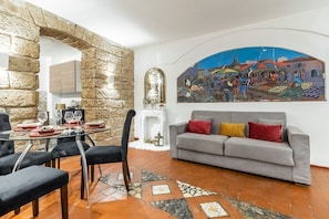 A dining area with a glass table, chairs, a stone wall, a grey sofa, and a colorful mural. - GuestHost - The Arches of Ballarò - Cortile Scimeca Apartment (Palermo)