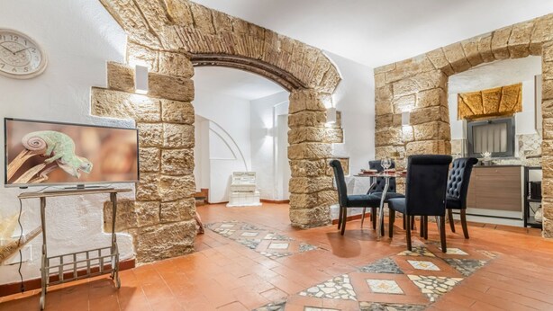 A room with stone arches, a dining area with chairs, a TV on a stand, and a tiled floor. - GuestHost - The Arches of Ballarò - Cortile Scimeca Apartment (Palermo)