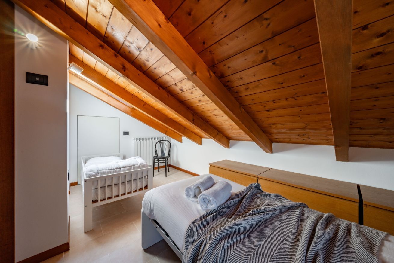 Une chambre avec un plafond en bois incliné, un berceau blanc, un lit recouvert d’une couverture à rayures et un banc en bois.