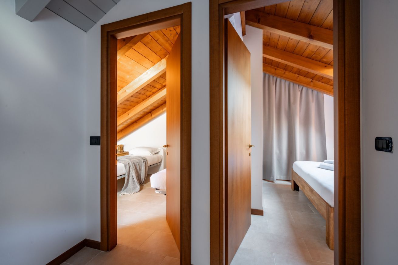 Ein Zimmer mit schrägem Dach, Holzbalken, einem Bett, einem Nachttisch und einem Fenster mit Vorhängen.