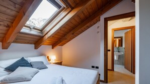 Ein Schlafzimmer mit Oberlicht, einem Bett mit Kissen und Handtüchern, einem Nachttisch mit Lampe und einem durchgehenden Türspalt sichtbaren Badezimmer.