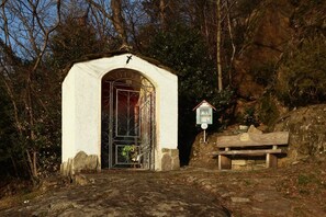 Una pequeña capilla blanca con puerta metálica y cruz en lo alto, rodeada de árboles y un banco de piedra cercano.