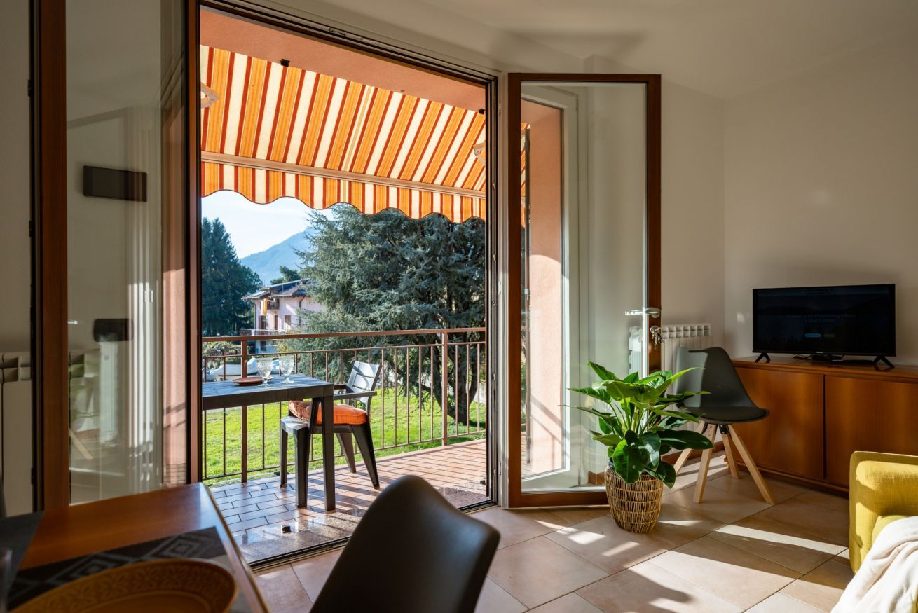 A modern living room with a sliding glass door leading to a balcony. There is a television on a wooden cabinet, a dining table with chairs, and a potted plant.