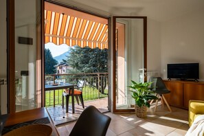 Een moderne woonkamer met een schuifglazen deur naar een balkon. Er staat een televisie op een houten kast, een eettafel met stoelen en een plant in een pot.