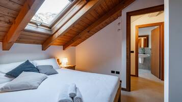 A bedroom with a skylight, a bed with pillows and towels, a nightstand with a lamp, and a bathroom visible through an open door.
