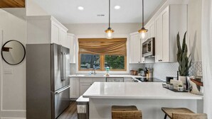 Una cocina moderna con armarios blancos, una isla grande con taburetes, electrodom ésticos de acero inoxidable e iluminación colgante.